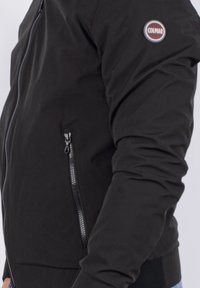 La giacca bomber nera presenta una chiusura con zip frontale, tasche laterali, polsini e orlo a costine, e una toppa rotonda con logo sul braccio sinistro.