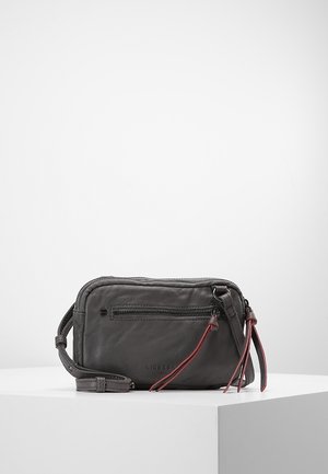 Mała ciemnoszara skórzana torebka crossbody z przednią kieszenią na zamek, regulowanym paskiem i czerwonymi skórzanymi uchwytami zamka na białej powierzchni ekspozycyjnej.