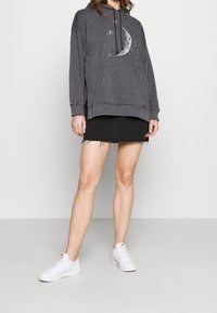 Sweat à capuche gris avec motif de lune, poche avant et capuche réglable par cordon. Porté avec une mini-jupe en denim noire et des baskets blanches.