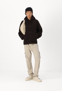 Hnědá fleecová mikina s přední kapsou, doplněná bežovými cargo kalhotami s bočními kapsami. Model má na sobě světle tenisky a bežovou crossbody kabelku.