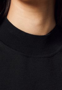 Schwarzer gestrickter Pullover mit einem gerippten, hohen Kragen. Der Stoff wirkt weich und glatt und hat eineeinfarbige Optik ohne sichtbare Muster oder Akzente.