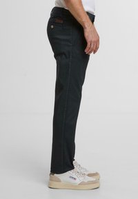 Schwarze Hosen mit schmaler Passform, strukturiertem Stoff, Seitentaschen und einem kleinen Lederpatch am Bund. Getragen mit weißen und beigen Sneakers.