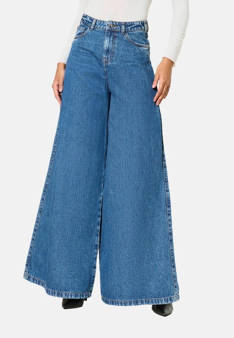 Model purtând blugi din denim albastru, cu talie înaltă și picioare largi, cu buzunare frontale și închidere cu nasturi, asortați cu pantofi negri cu vârf ascuțit.