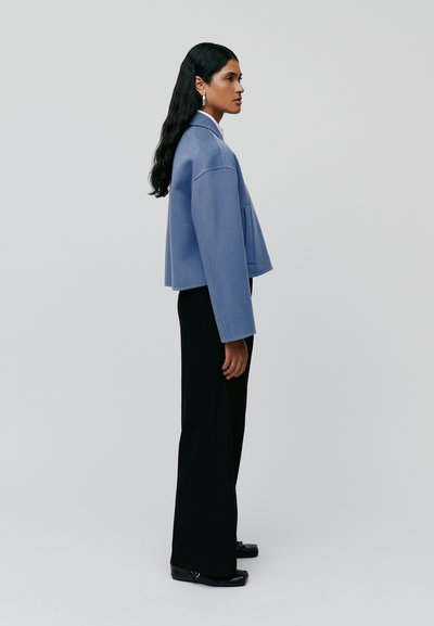 Giacca cropped blu chiaro con maniche larghe e una tasca, abbinata a pantaloni neri a gamba larga e scarpe nere.