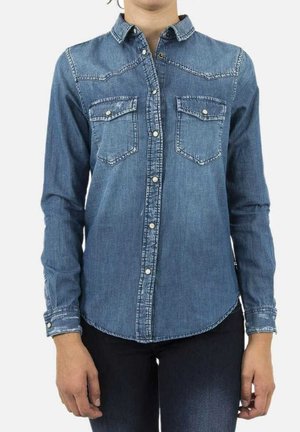 Osoba ubrana w niebieską jeansową koszulę na guziki z dwoma kieszeniami na piersi i długimi rękawami, stojąca na jednolitym tle.