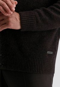 Handen die de manchet van een donkerbruine wollen cardigan met knopen en een klein rechthoekig merklabel nabij de onderzoom aanpassen.