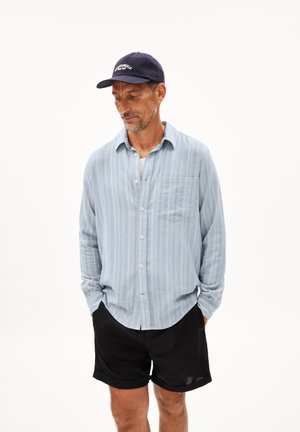 Light blue striped button-up shirt with a pocket, worn over a white undershirt, paired with black shorts and a navy cap. Texture is soft.
