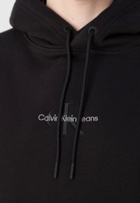 Sweat à capuche noir avec broderie du logo Calvin Klein Jeans sur le devant et cordons noirs, porté par une personne visible du cou vers le bas.