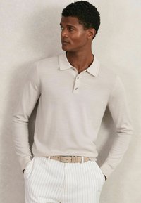 Beige Langarm-Polohemd mit Kragen, ausgestattet mit drei Knöpfen und einer weichen Textur, kombiniert mit weißen gestreiften Hosen und einem hellen Gürtel.