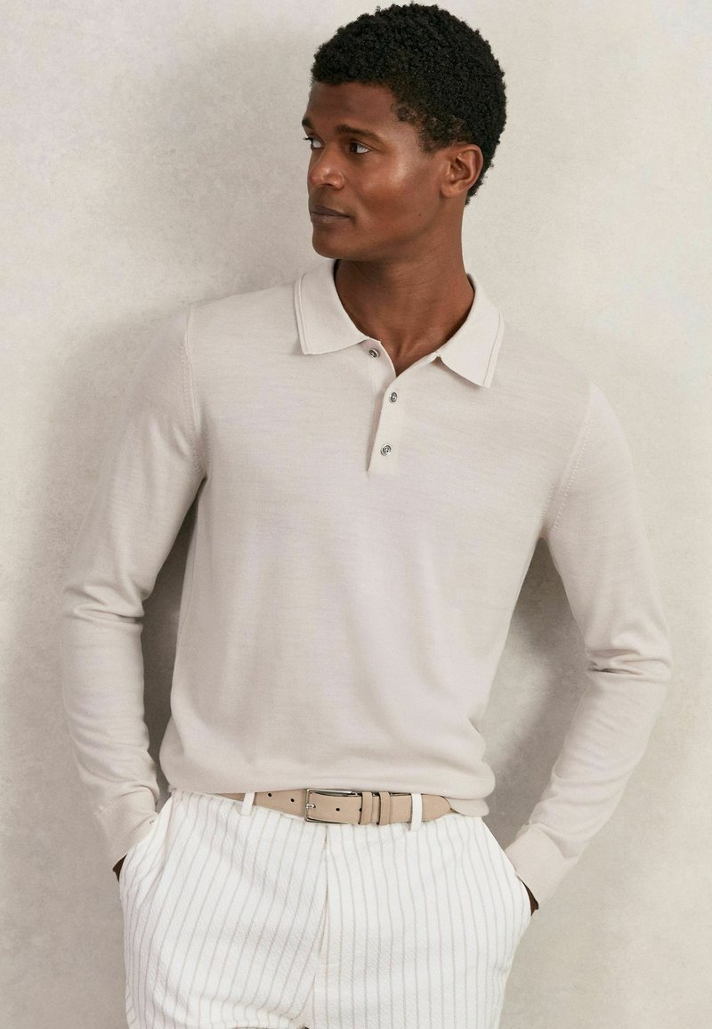 Beige Langarm-Polohemd mit Kragen, ausgestattet mit drei Knöpfen und einer weichen Textur, kombiniert mit weißen gestreiften Hosen und einem hellen Gürtel.
