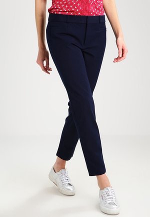 Kobieta ubrana w granatowe spodnie o dopasowanym kroju, białe sneakersy i czerwoną bezrękawnikową bluzkę z nadrukiem ptaków, stojąca na tle jednolitego tła.