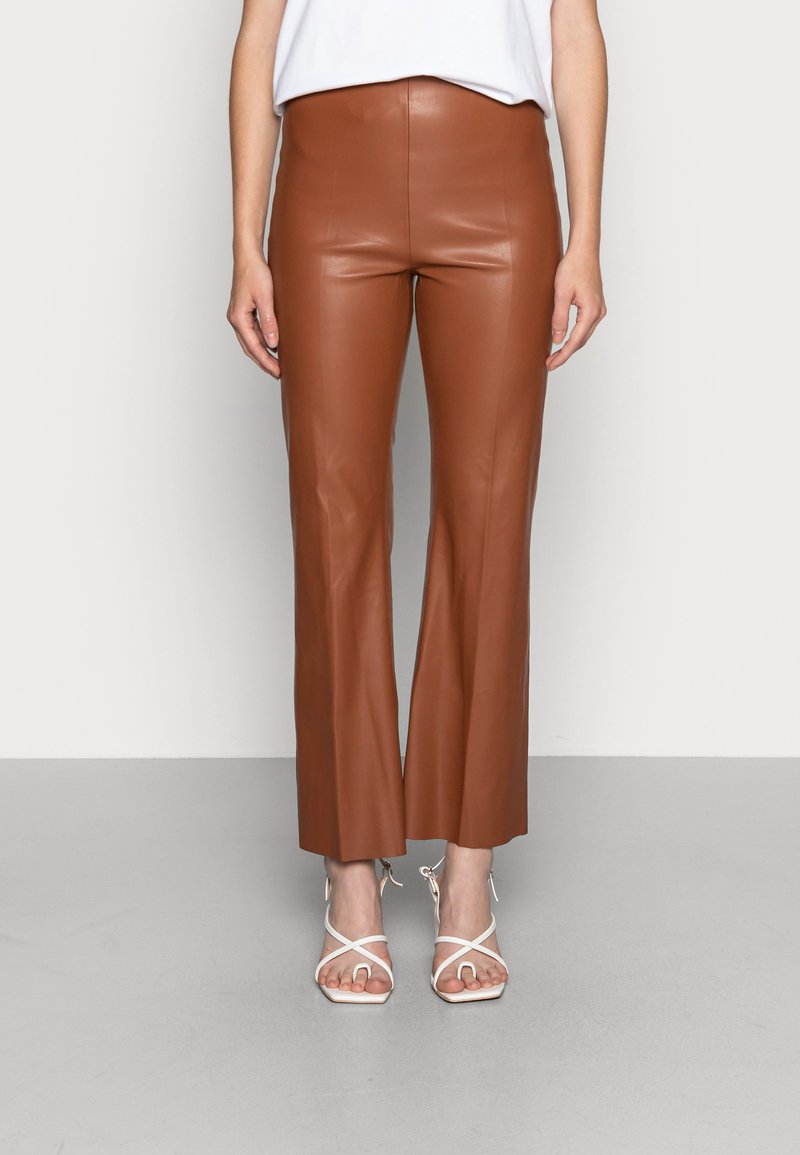 Bruine faux leren flared broek met een hoge taille, een gladde textuur en een strakke uitstraling. Draagt met witte gespsandalen met hakken.