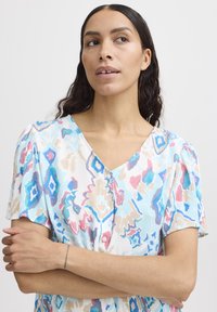 Mehrfarbige Bluse mit V-Ausschnitt, kurzen Puffärmeln und einem dekorativen Muster in Blau-, Pink- und Beigetönen, hergestellt aus leichtem Stoff.