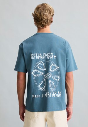 Person mit kurzen blonden Haaren, die ein blaues T-Shirt mit weißem Austernparty-Grafik und Text auf der Rückseite trägt.