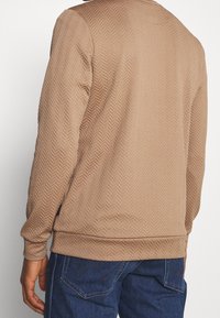 Felpa marrone con texture e motivo a spina di pesce, con polsini e orlo a costine, vestibilità rilassata, indossata sopra jeans blu. Visualizzazione della parte posteriore.