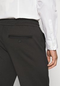 Schwarze Hose mit glatter Textur, elastischem Bund und einer Gesäßtasche mit Knopf. Getragen mit einem weißen Hemd mit Knöpfen.