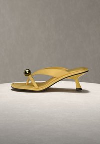 Κίτρινο πέδιλο με τακούνι και λουράκι με δάχτυλο, διακοσμημένο με στρογγυλή μεταλλική χρυσή λεπτομέρεια, σε ουδέτερο φόντο και επιφάνεια.