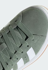 Zelené semišové tenisky s bielymi koženými pruhmi, textúrovanými šnúrkami a gumovou podrážkou. Majú zaoblenú špičku a detaily šitia.