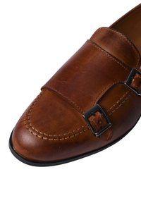 Mocassino in pelle marrone con due fibbie nere, dettagli cuciti, punta arrotondata e texture liscia. La suola in gomma offre aderenza.