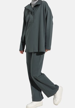 Person trägt lockeres dunkelgraues Langarmshirt und Hose mit grauem Kopftuch und weißen Turnschuhen, steht auf einem Fuß.