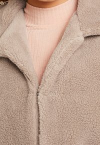 Lys grå, struktureret fleecejakke med reverskrave og frontlynlås. Båret over en blød, ribbet, lys rosa top.