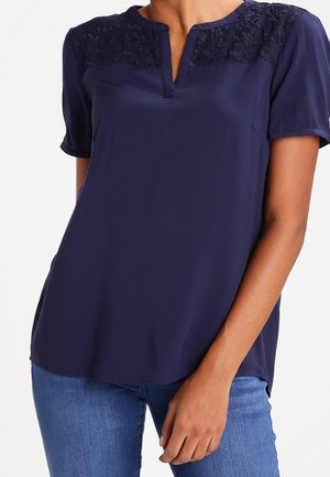 Marineblå bluse med korte ærmer med blonde detaljer på skuldrene og hakket halsudskæring, båret med blå jeans.