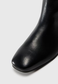 Primer plano de la punta y el costado de una bota de tobillo de cuero negro con un acabado suave y ligeramente texturizado sobre un fondo blanco.