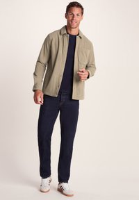 Man smiling, wearing beige corduroy jacket over navy shirt, dark jeans, and white sneakers, standing against plain light background.