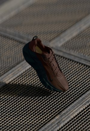 Brązowy skórzany sneaker z czarną podeszwą i sznurowadłami unoszący się nad teksturowanym metalowym rusztem.