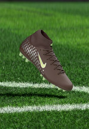 Καφέ ποδοσφαιρικό παπούτσι με ασημένιες κουκκίδες και κίτρινο λογότυπο Nike, δεμένο με κορδόνια, αιωρείται πάνω από πράσινο χορτάρι κοντά σε λευκές γραμμές γηπέδου.