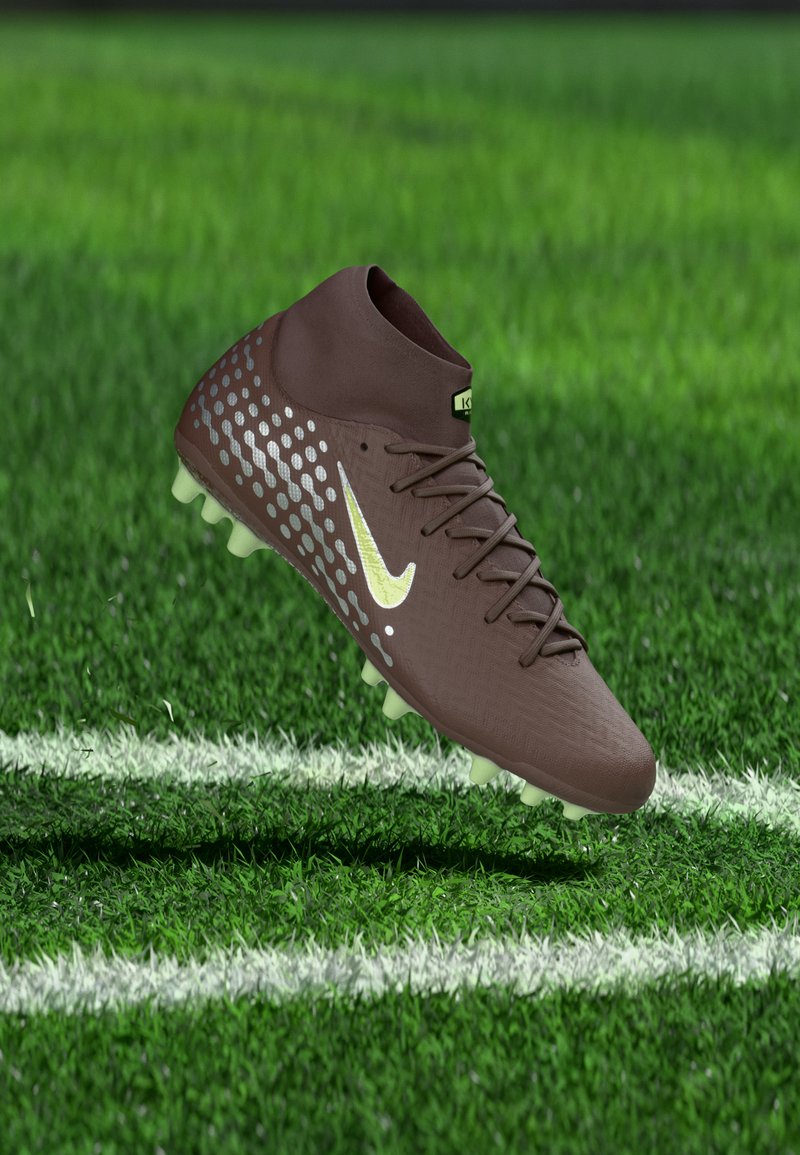 Καφέ ποδοσφαιρικό παπούτσι με ασημένιες κουκκίδες και κίτρινο λογότυπο Nike, δεμένο με κορδόνια, αιωρείται πάνω από πράσινο χορτάρι κοντά σε λευκές γραμμές γηπέδου.