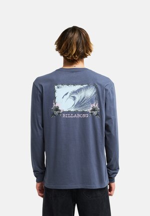 Persoon met kort bruin haar draagt een blauw shirt met lange mouwen met een grote golf en bloemenafbeelding en het woord "BILLABONG" op de rug.