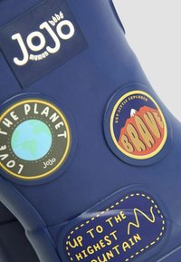 Bolsa de goma azul marino con parches circulares: "Ama el Planeta", "Valiente" y "Hasta la Montaña Más Alta". Textura suave. Logo visible.