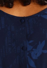 Woman wearing a dark blue blouse with subtle leaf and geometric patterns and fabric-covered buttons along the front.