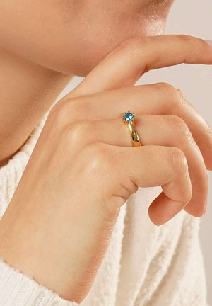 Gold ring featuring a round, light blue gemstone at the center, nestled in a six-prong setting, worn on a hand with neutral skin tone.