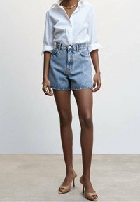Camisa blanca de botones metida dentro de unos pantalones cortos de denim de cintura alta, color azul clásico, textura suave, combinada con sandalias de tacón alto en color nude.