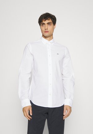 Man die een wit overhemd met knopen en kraag draagt en een zwarte broek, staand tegen een effen witte achtergrond.