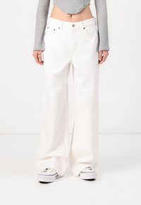 Jeans blancs à jambes larges en denim, avec une taille haute et un design à cinq poches standard, associés à un haut gris à manches longues et des baskets.