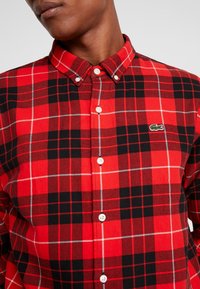Camisa de cuadros en rojo y negro con patrón de cuadros blancos, cuello abotonado y un pequeño logo verde en el pecho. Hecha de tela tejida.