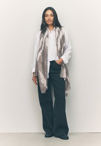 Sciarpa leggera con motivo grigio e bianco drappeggiata su una camicia bianca, abbinata a jeans ampi scuri e scarpe nere.