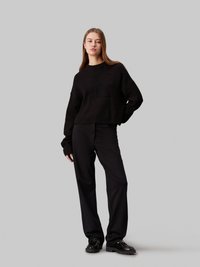 Maglione nero a maglia con collo rotondo e texture bulky, abbinato a pantaloni neri a vestibilità morbida e scarpe nere lucide.