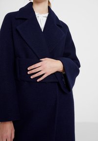 Marineblauer Wollmantel mit tailliertem Gürtel, Reverskragen und weiten Ärmeln. Die Textur ist weich und hat eine gebürstete Oberfläche für zusätzliche Wärme.
