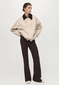 Veste bomber beige avec un col noir, coupe oversize et texture lisse, associée à un pantalon évasé marron foncé et des chaussures crème.