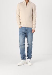 Pull beige texturé à fermeture éclair sur le devant jusqu'au quart, associé à un jean bleu clair et des baskets blanches. Design simple et décontracté avec des poignets côtes.