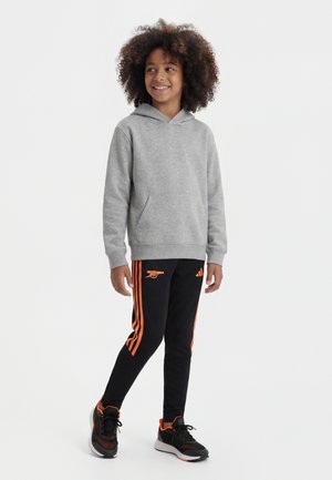 Kind mit lockigem Haar, das einen grauen Hoodie, schwarze Sporthosen mit orangen Streifen und schwarze Sportschuhe trägt, steht und lächelt.