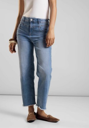 Mujer con jeans cropped azul claro, top sin mangas blanco, zapatos marrones tipo slip-on y pulseras en la muñeca izquierda, de pie contra un fondo gris.