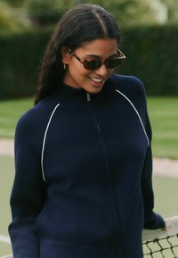 Pull en maille bleu marine à fermeture éclair avec passepoil blanc le long des épaules, fabriqué en tissu doux. Le mannequin porte des lunettes de soleil écaille de tortue et a les cheveux longs et foncés.