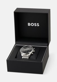 Montre en acier inoxydable argenté avec un cadran noir, fonctionnalités chronographe, présentée dans une boîte noire avec le logo "BOSS".