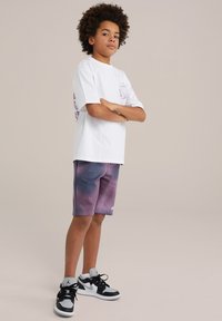 Biela tričko s fialovým grafickým motívom na ľavej strane, v kombinácii s šortkami s fialovo-modrým gradientom a bodkovaným vzorom. Čierno-biele tenisky.