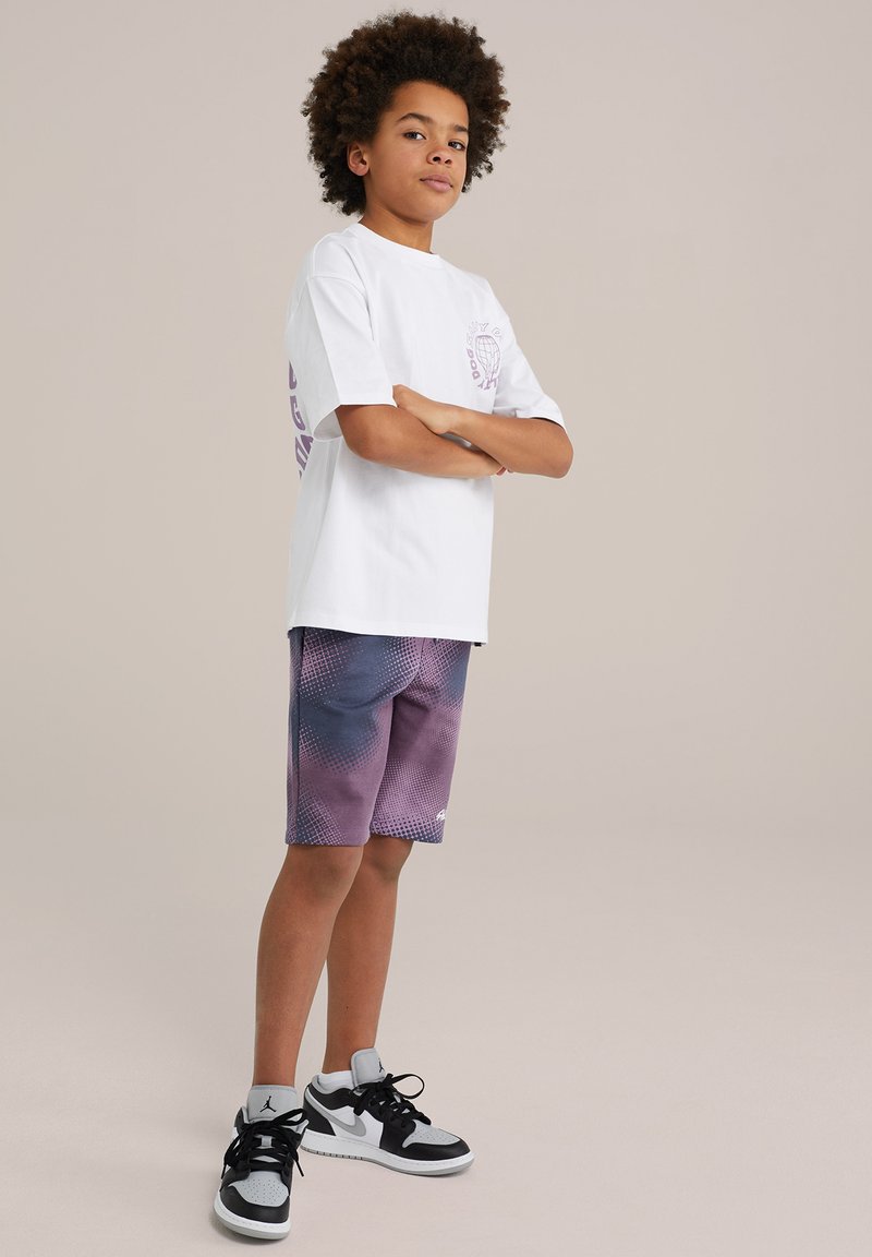 Biela tričko s fialovým grafickým motívom na ľavej strane, v kombinácii s šortkami s fialovo-modrým gradientom a bodkovaným vzorom. Čierno-biele tenisky.